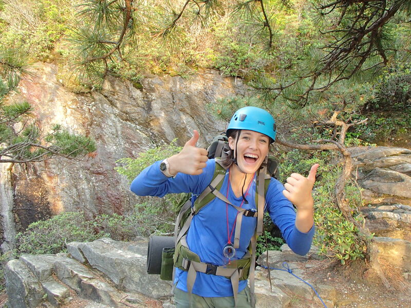 A woman wearing a blue helmet and a blue long-sleeved shirt is standing outdoors, giving two thumbs up. She has a backpack on and a watch on her left wrist. The background features a rocky cliff face with greenery and trees. The woman appears to be happy and energetic, possibly on a hiking or climbing adventure.